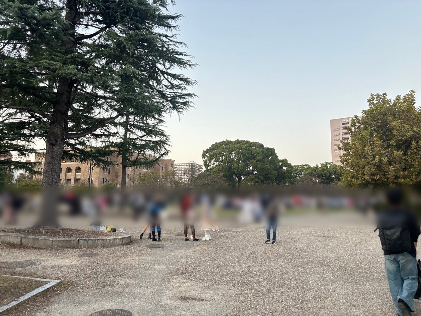A park in Nagoya at dusk with tall trees, an old brick building, and many people in colorful costumes (faces blurred)