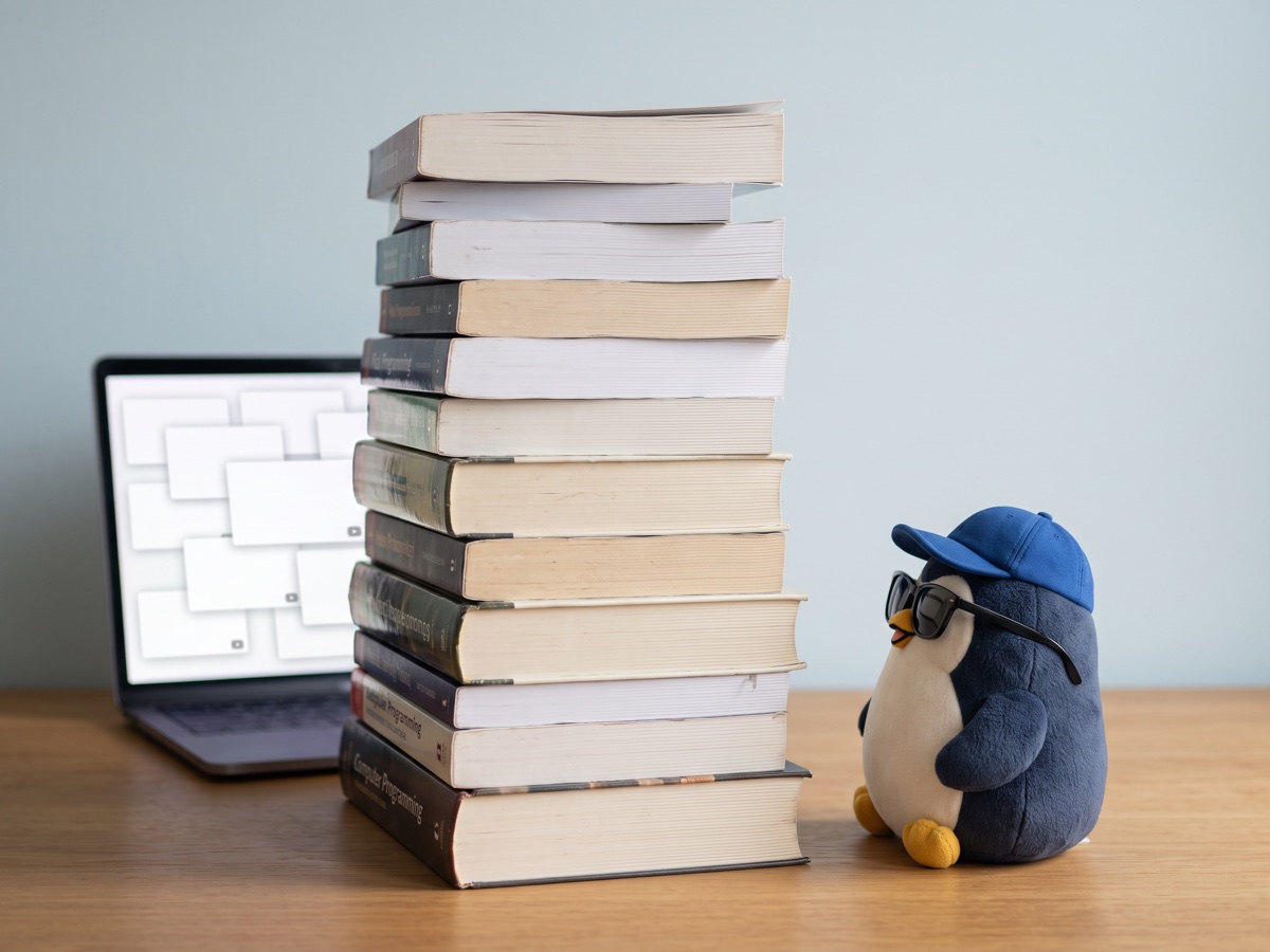 A small navy plush penguin with a blue cap and sunglasses looks up at a tall stack of programming books next to a laptop